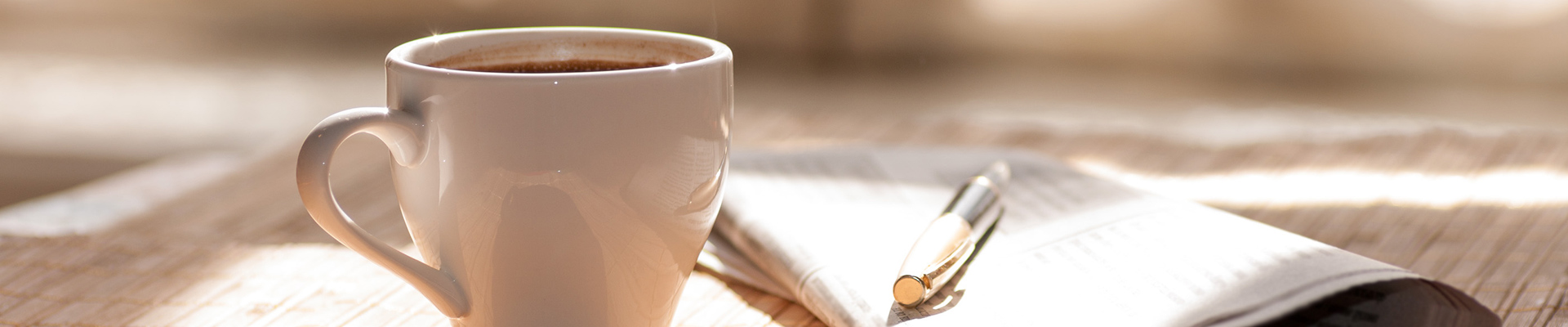 White ceramic coffee mug on a sunlit wooden table with a notebook and pen in the background, creating a cozy workspace scene.