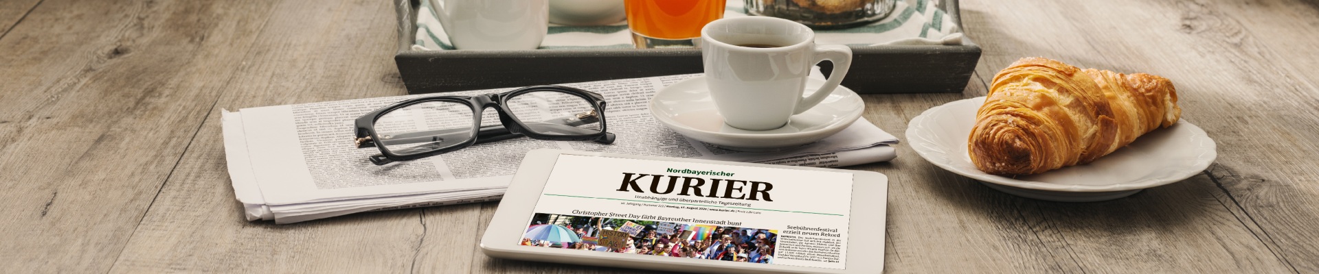 Tablet showing a German newspaper beside a pair of glasses on a wooden table with a cup of coffee and a croissant on a plate.