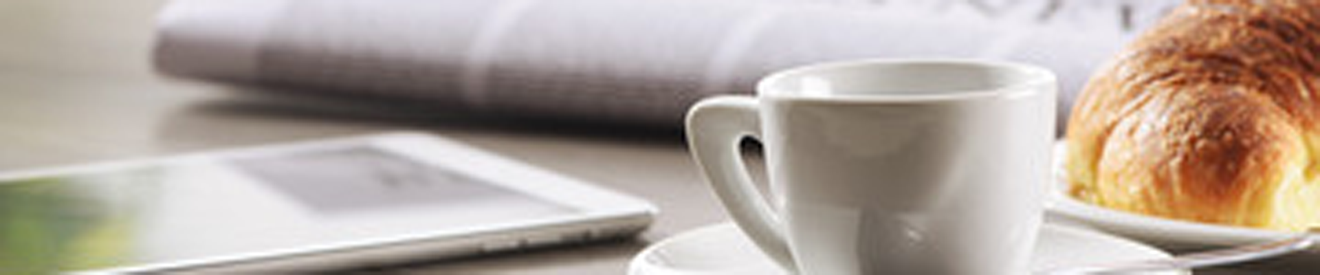 White coffee cup on a saucer with a flaky croissant and a tablet on a light breakfast table